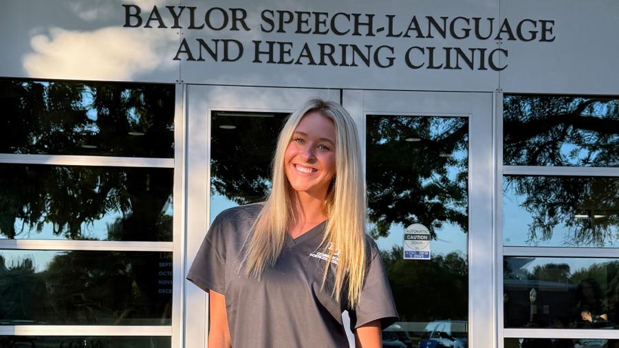 Taylor Field stands in front of the Baylor Speech-Language and Hearing Clinic, while wearing scrubs.